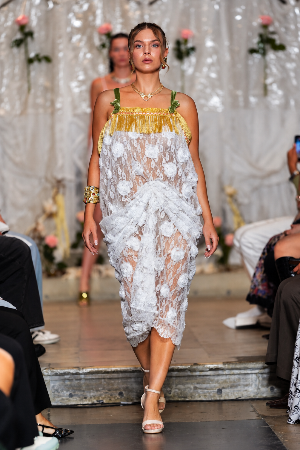 A model walks a fashion runway wearing a sheer white floral lace dress with a gold fringe top and green shoulder straps, set against a decorated white draped backdrop with flowers.