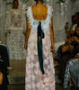 A model viewed from behind on a runway wearing a long, textured white and pale pink dress with a large dark green ribbon tied at the open back. The runway show is in a real environment with seated spectators visible on both sides.