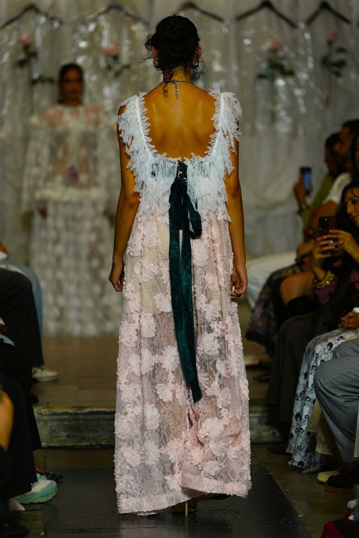 A model viewed from behind on a runway wearing a long, textured white and pale pink dress with a large dark green ribbon tied at the open back. The runway show is in a real environment with seated spectators visible on both sides.