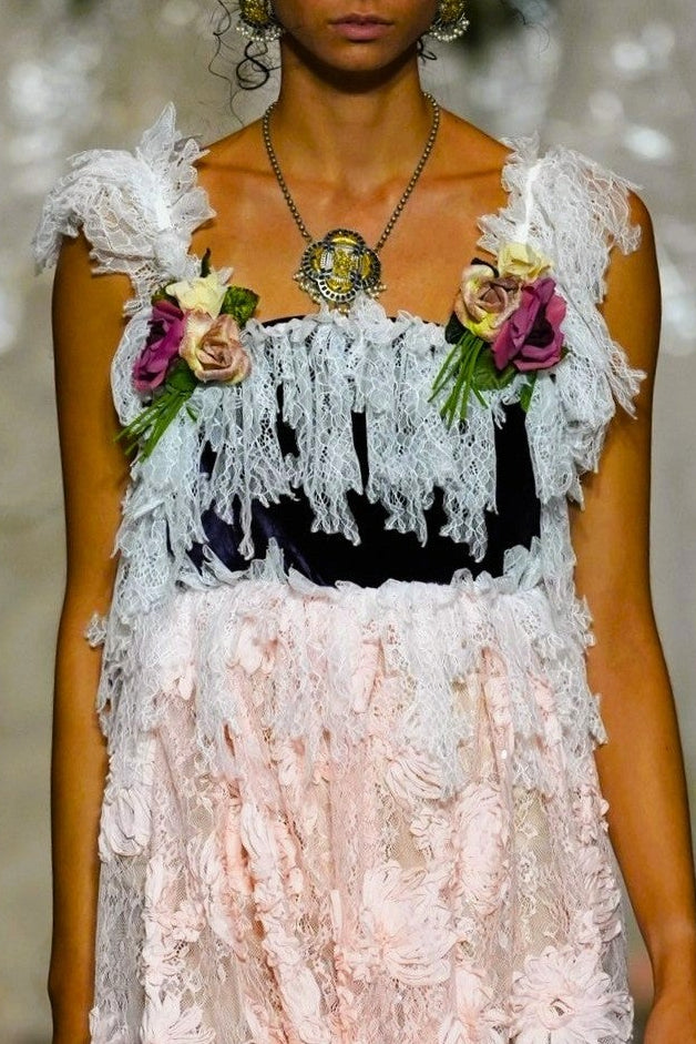 A model walks the runway wearing a sleeveless maxi dress with a black bodice, white floral applique, and a long sheer white skirt with large floral patterns.