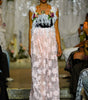 A model walks the runway wearing a sleeveless maxi dress with a black bodice, white floral applique, and a long sheer white skirt with large floral patterns.