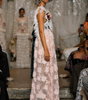 A model walks a fashion runway wearing a floor-length, sleeveless pale pink lace gown with 3D floral appliqué, a dark teal sash, and a floral chest bouquet, with a white draped backdrop and seated audience members in the foreground.