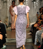 A model from behind wearing a lavender floral print midi dress with light pink ruffled sleeves, walking down a fashion runway.