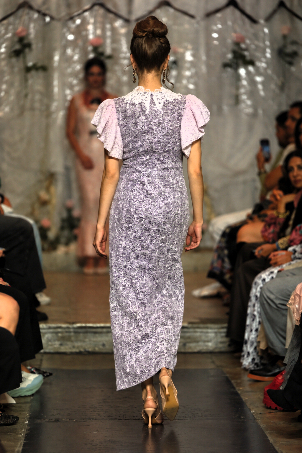 A model from behind wearing a lavender floral print midi dress with light pink ruffled sleeves, walking down a fashion runway.