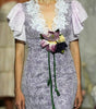 A model walks a fashion runway wearing a lavender and white patterned midi dress with ruffled sleeves, a lace V-neckline, and a purple satin inner slit. The runway is surrounded by seated spectators and a blurred model in the background.