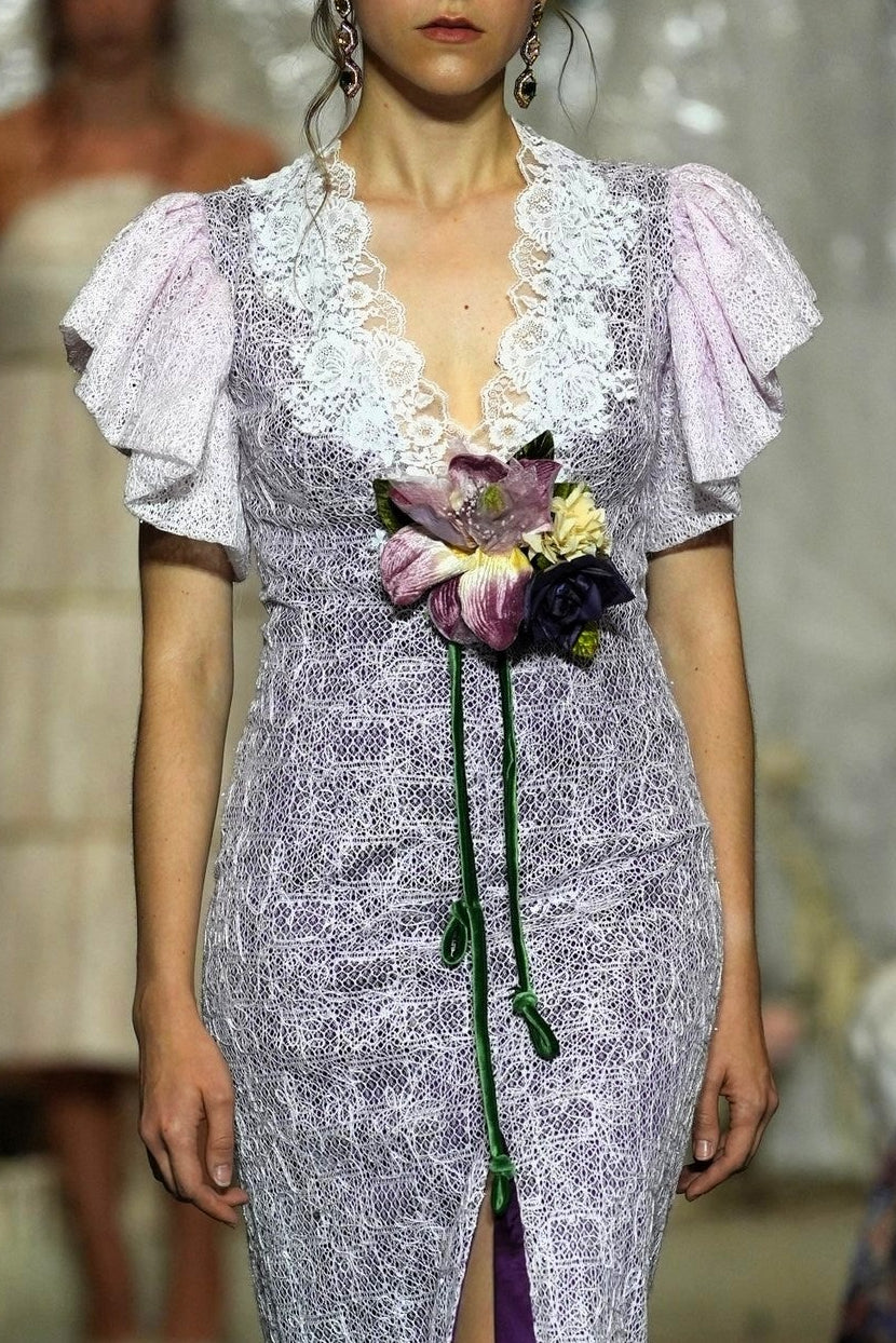 A model walks a fashion runway wearing a lavender and white patterned midi dress with ruffled sleeves, a lace V-neckline, and a purple satin inner slit. The runway is surrounded by seated spectators and a blurred model in the background.