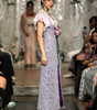 A model walks a fashion runway wearing a lavender and white patterned maxi dress with ruffled sleeves, a purple side panel, and holding a pink flower bouquet.