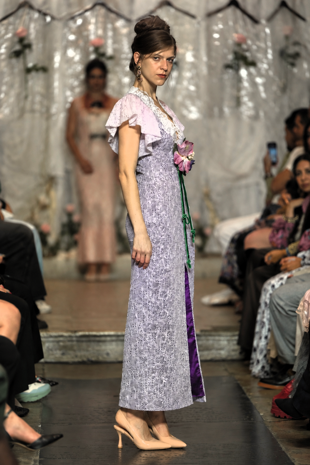 A model walks a fashion runway wearing a lavender and white patterned maxi dress with ruffled sleeves, a purple side panel, and holding a pink flower bouquet.