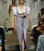 A model walks a fashion runway wearing a lavender and white patterned midi dress with ruffled sleeves, a lace V-neckline, and a purple satin inner slit. The runway is surrounded by seated spectators and a blurred model in the background.