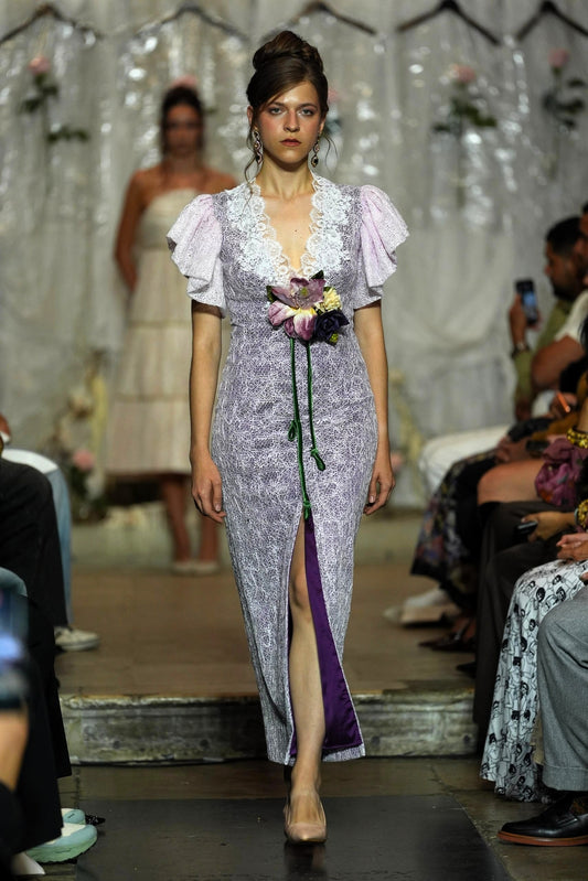 A model walks a fashion runway wearing a lavender and white patterned midi dress with ruffled sleeves, a lace V-neckline, and a purple satin inner slit. The runway is surrounded by seated spectators and a blurred model in the background.