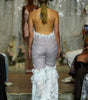 A model viewed from behind on a runway wearing a strapless lace jumpsuit with a high open back and a voluminous ruffled white lace hem. The background shows a fashion show setting with other models and audience members.