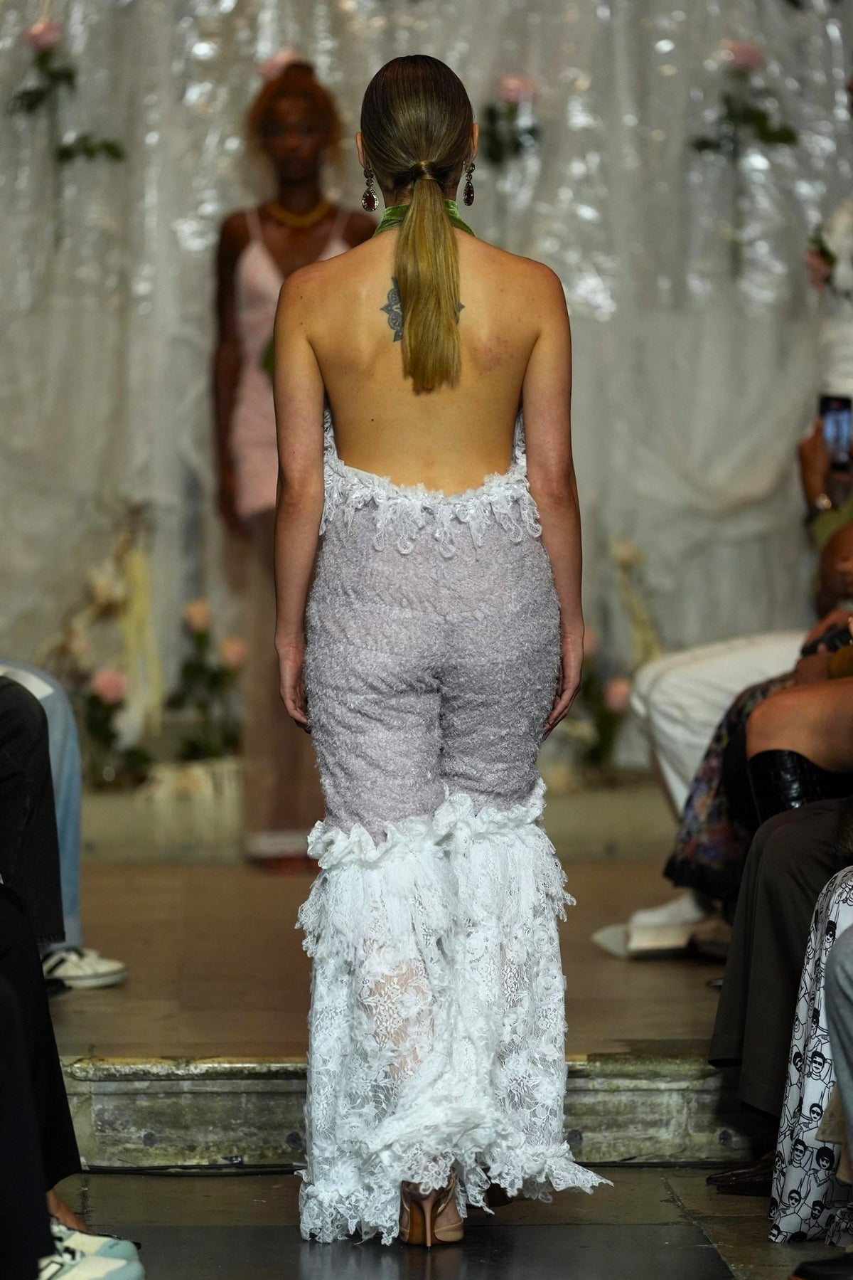 A model viewed from behind on a runway wearing a strapless lace jumpsuit with a high open back and a voluminous ruffled white lace hem. The background shows a fashion show setting with other models and audience members.