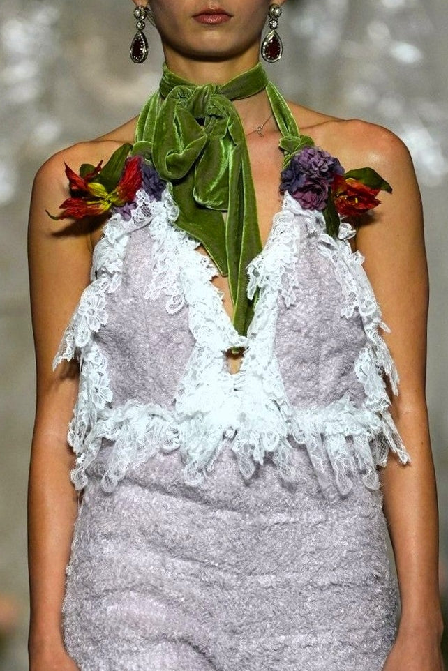 A model walks the runway wearing a textured lavender and white halter jumpsuit with lace ruffle trim and a green neck scarf with floral accents.