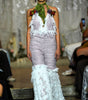 A model walks the runway wearing a textured lavender and white halter jumpsuit with lace ruffle trim and a green neck scarf with floral accents.