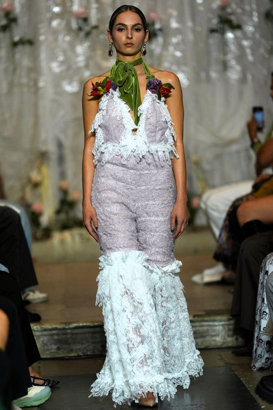 A model walks the runway wearing a textured lavender and white halter jumpsuit with lace ruffle trim and a green neck scarf with floral accents.