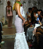 A model walks down a fashion runway wearing a sleeveless, backless mermaid-style gown with a pale lavender textured bodice and a white ruffled mermaid skirt. She is accessorized with a green scarf and a red rose at the neckline.