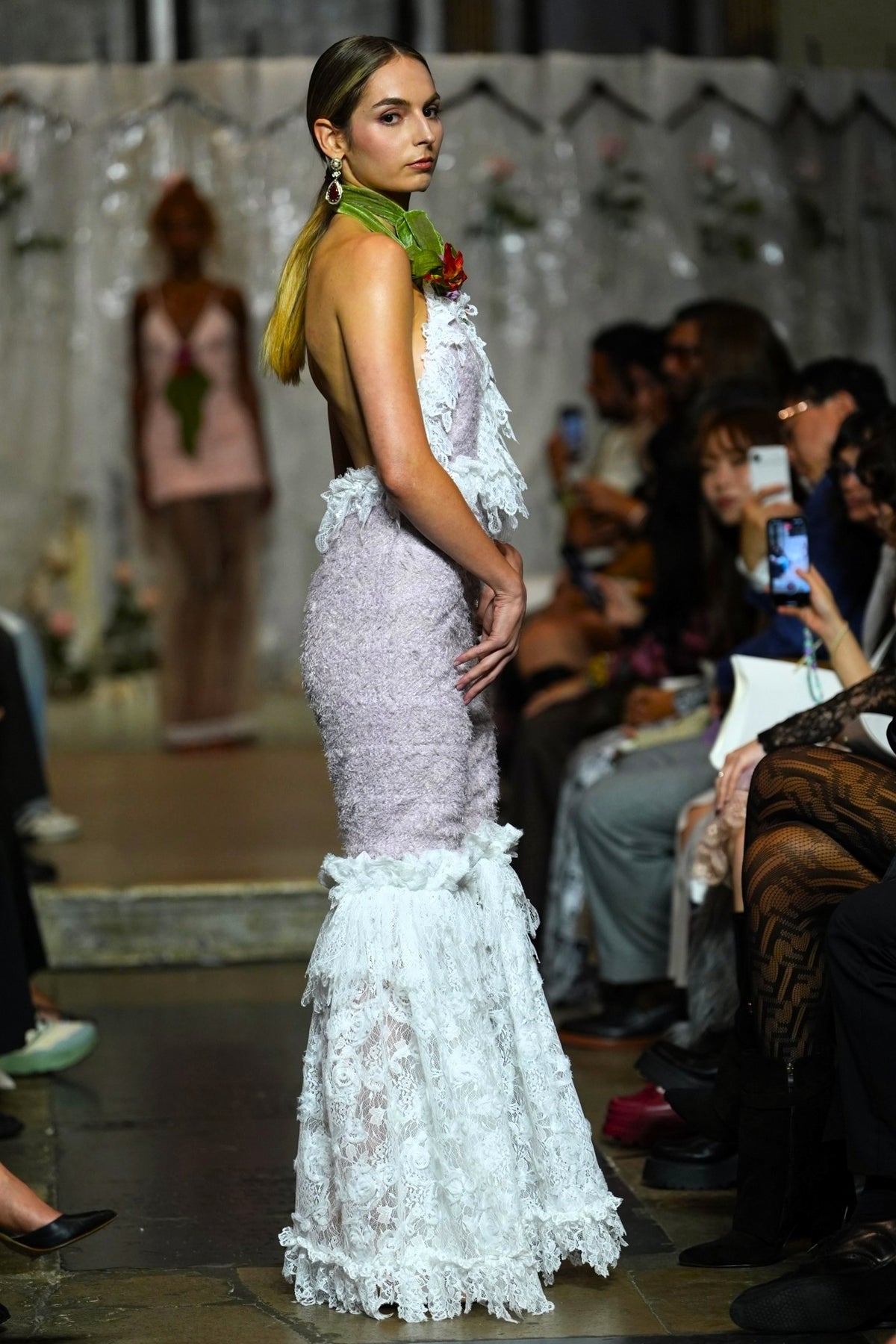 A model walks down a fashion runway wearing a sleeveless, backless mermaid-style gown with a pale lavender textured bodice and a white ruffled mermaid skirt. She is accessorized with a green scarf and a red rose at the neckline.