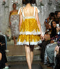 A model from behind wearing a yellow and white tiered lace dress on a fashion runway, with seated spectators visible on both sides.
