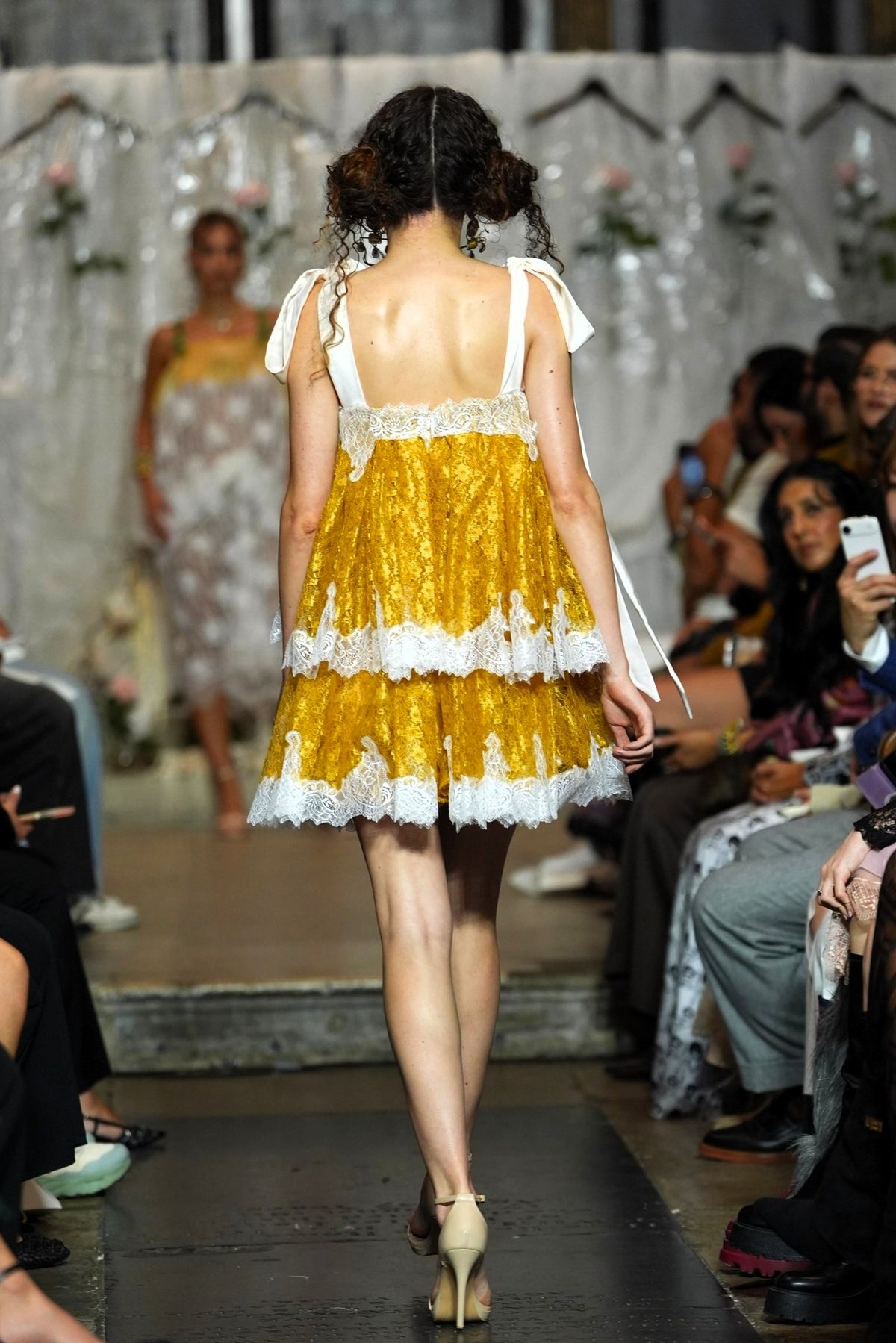 A model from behind wearing a yellow and white tiered lace dress on a fashion runway, with seated spectators visible on both sides.