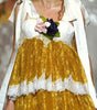 A model walks the runway wearing a short white dress with a voluminous gold skirt, white lace trim, and a white draped cape. She holds a small bouquet of pink and dark flowers.