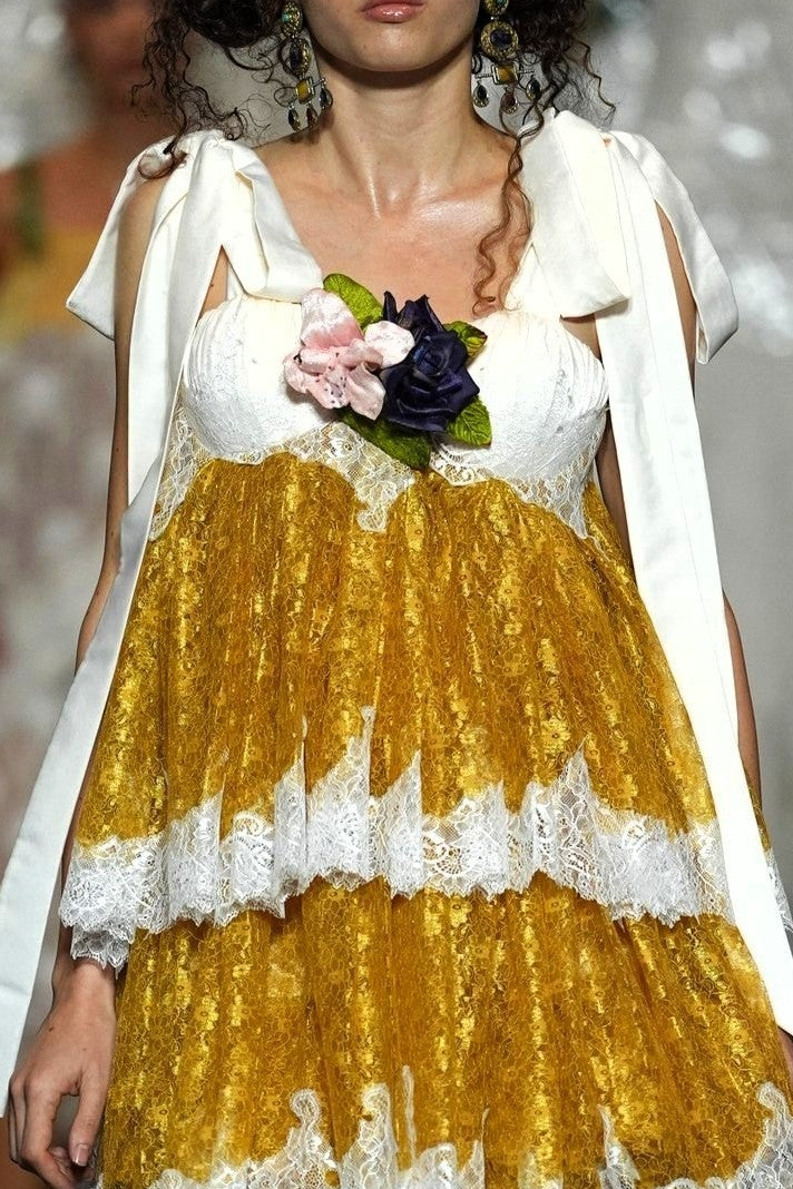 A model walks the runway wearing a short white dress with a voluminous gold skirt, white lace trim, and a white draped cape. She holds a small bouquet of pink and dark flowers.