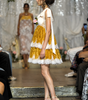 A model walks a fashion runway wearing a short white dress with a tiered skirt in shiny gold and white lace, paired with beige high heels. The runway is decorated with white drapes and flowers, with audience members seated on both sides.