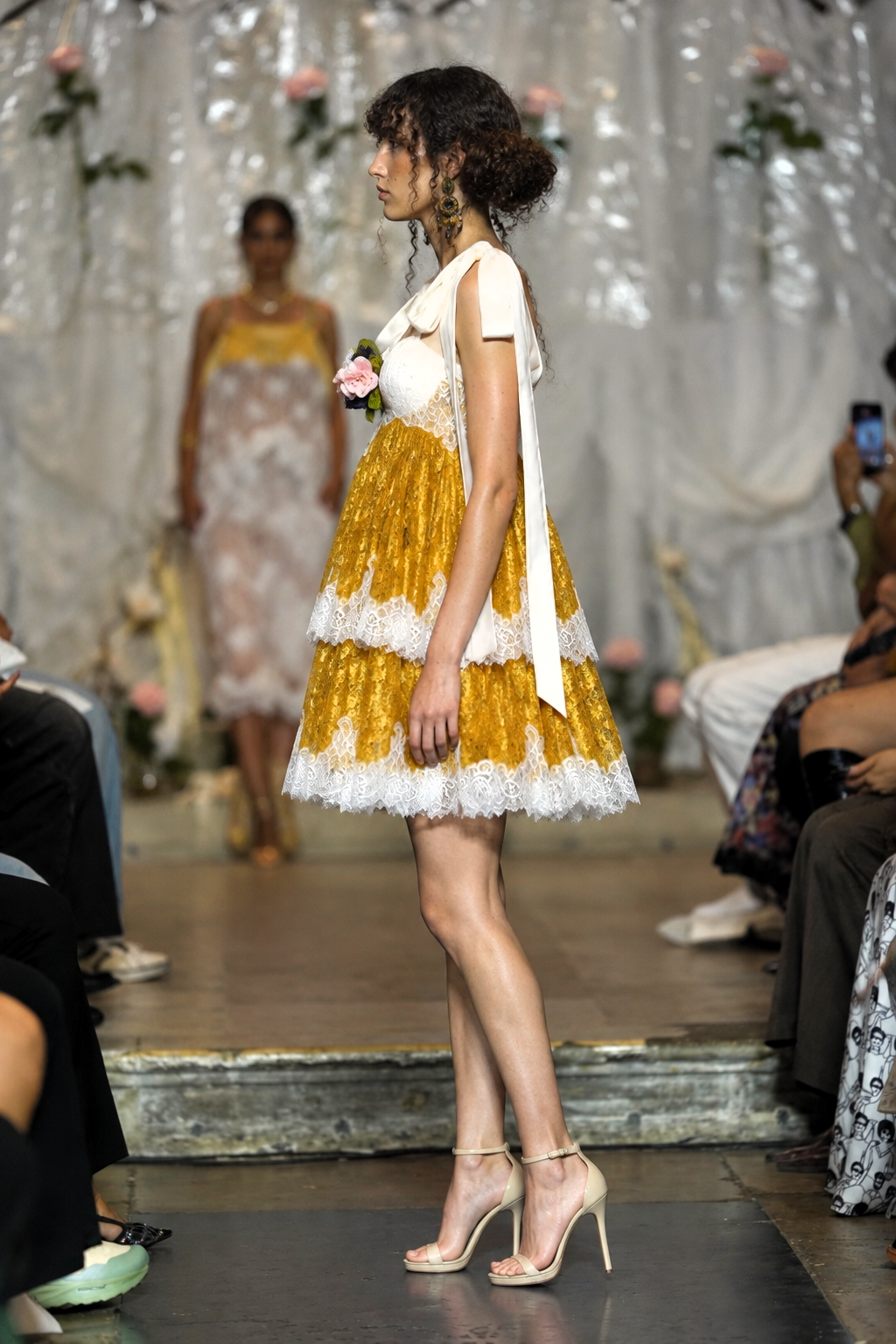 A model walks a fashion runway wearing a short white dress with a tiered skirt in shiny gold and white lace, paired with beige high heels. The runway is decorated with white drapes and flowers, with audience members seated on both sides.