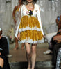 A model walks the runway wearing a short white dress with a voluminous gold skirt, white lace trim, and a white draped cape. She holds a small bouquet of pink and dark flowers.