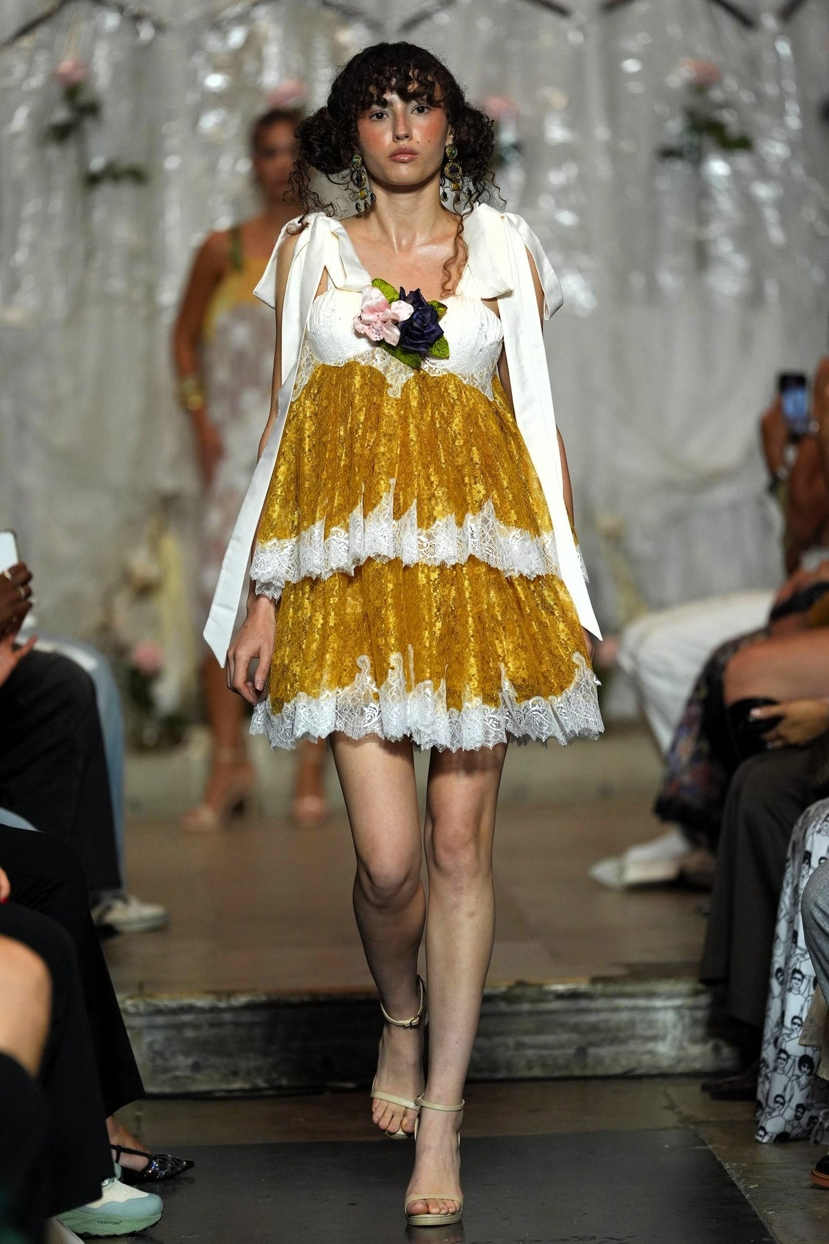 A model walks the runway wearing a short white dress with a voluminous gold skirt, white lace trim, and a white draped cape. She holds a small bouquet of pink and dark flowers.