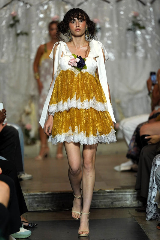 A model walks the runway wearing a short white dress with a voluminous gold skirt, white lace trim, and a white draped cape. She holds a small bouquet of pink and dark flowers.