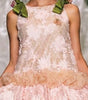 A model walks a fashion runway wearing a sleeveless light pink floral lace dress with a ruffled tulle waistband and matching pink shoes.