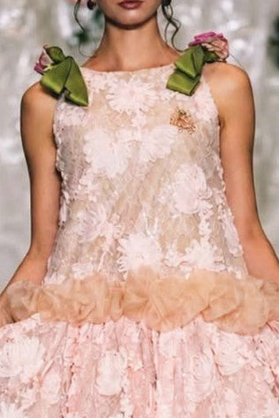 A model walks a fashion runway wearing a sleeveless light pink floral lace dress with a ruffled tulle waistband and matching pink shoes.