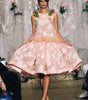 A model walks a fashion runway wearing a sleeveless light pink floral lace dress with a ruffled tulle waistband and matching pink shoes.