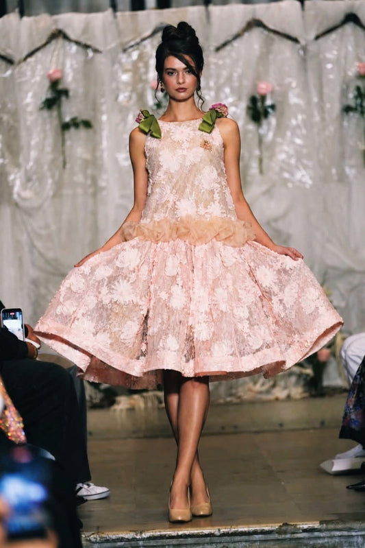 A model walks a fashion runway wearing a sleeveless light pink floral lace dress with a ruffled tulle waistband and matching pink shoes.