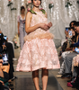 A model walks the runway wearing a light pink floral lace dress with a feathered waistband and beige heels, with audience members seated on both sides of the catwalk.