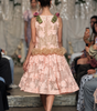 A model walks down a fashion runway wearing a sleeveless light pink lace dress with floral applique, a ruffled waist, and pink fabric flowers with green ribbon accents on the shoulders.