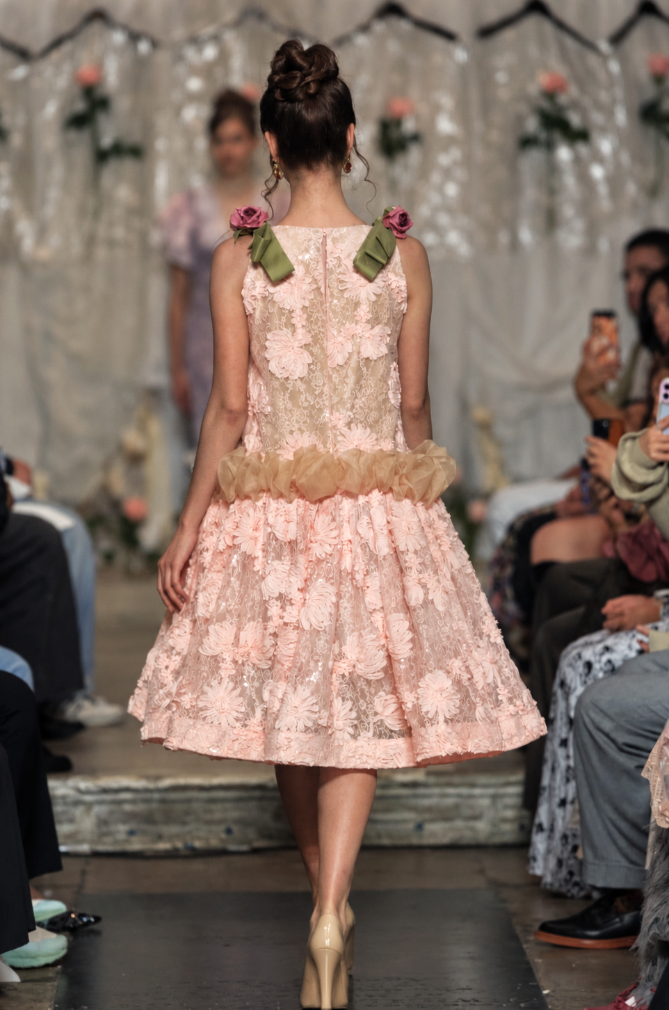 A model walks down a fashion runway wearing a sleeveless light pink lace dress with floral applique, a ruffled waist, and pink fabric flowers with green ribbon accents on the shoulders.