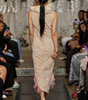 A model viewed from behind walking down a runway wearing a sleeveless, textured beige maxi dress with a high ruffled collar and pink fringe at the hem, paired with beige high heels.