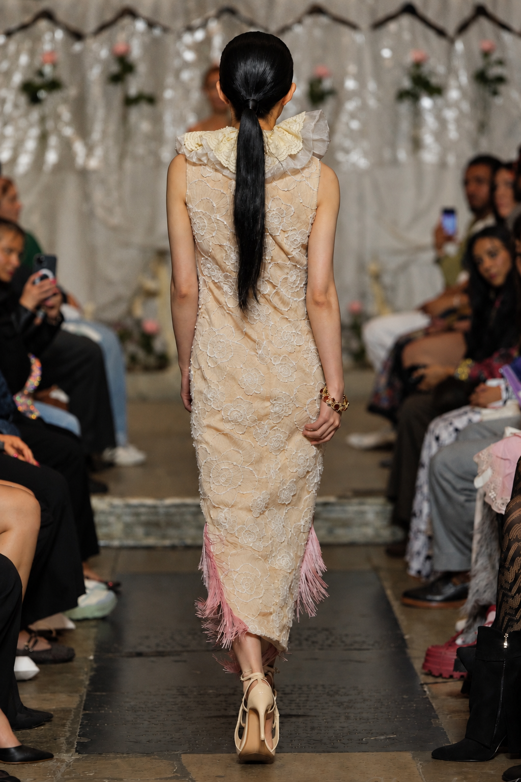 A model viewed from behind walking down a runway wearing a sleeveless, textured beige maxi dress with a high ruffled collar and pink fringe at the hem, paired with beige high heels.