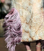 A model walks the runway wearing a sleeveless beige lace maxi dress with a large white ruffled collar, a pink bow, and pink feather trim at the hem.