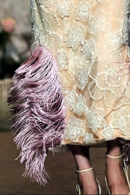 A model walks the runway wearing a sleeveless beige lace maxi dress with a large white ruffled collar, a pink bow, and pink feather trim at the hem.