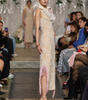 A model walks the runway wearing a sleeveless cream lace dress with pink fringe details and a high ruffled collar, photographed at a fashion show with seated spectators on both sides.