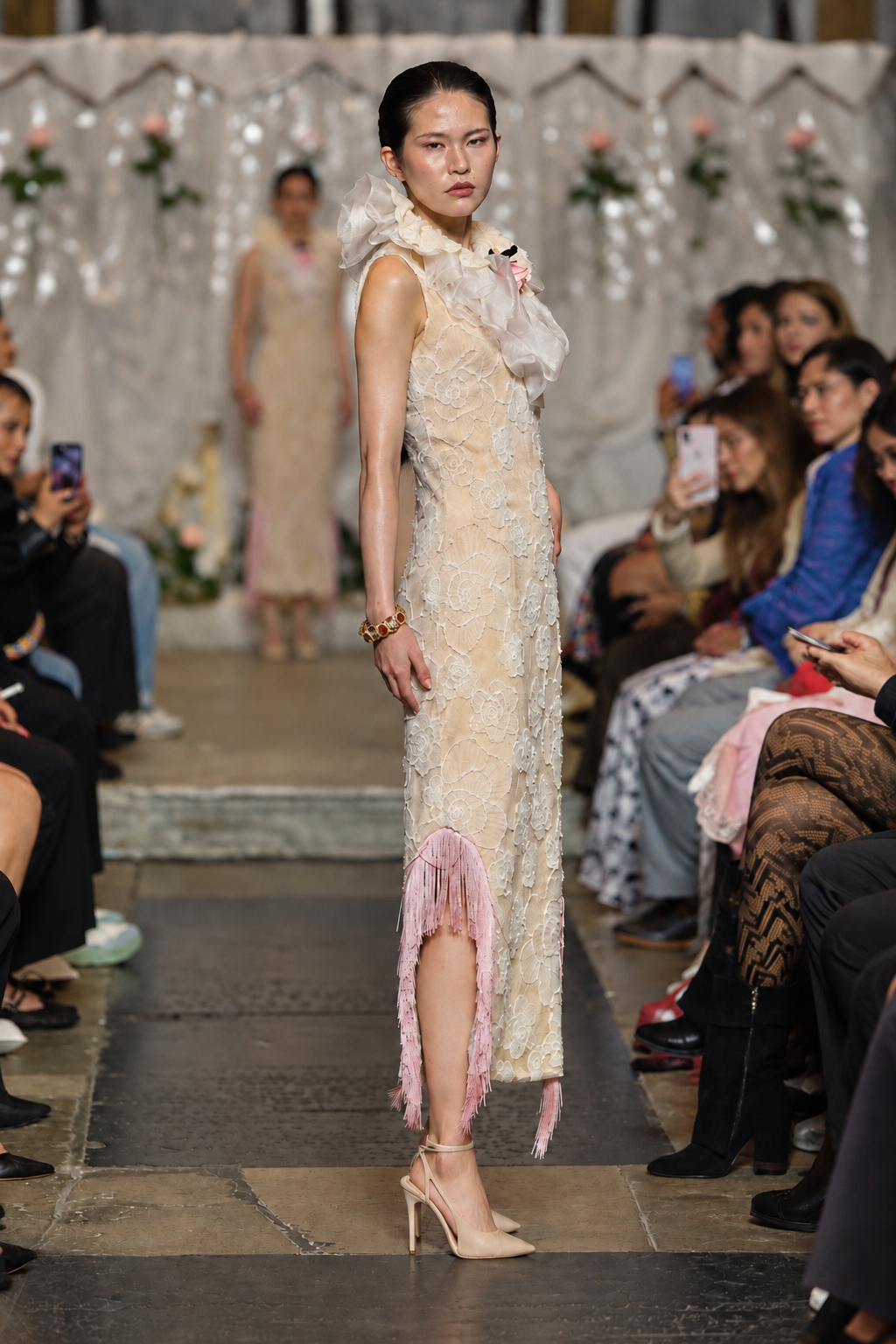 A model walks the runway wearing a sleeveless cream lace dress with pink fringe details and a high ruffled collar, photographed at a fashion show with seated spectators on both sides.