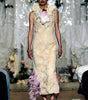A model walks the runway wearing a sleeveless beige lace maxi dress with a large white ruffled collar, a pink bow, and pink feather trim at the hem.