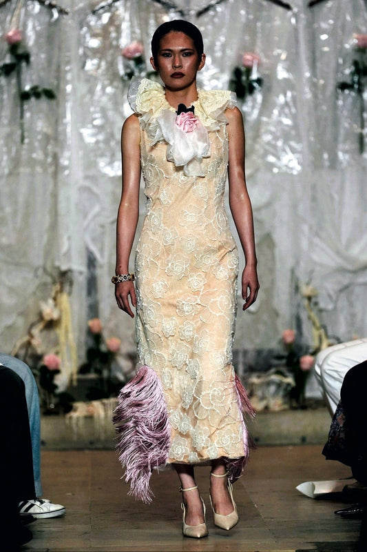A model walks the runway wearing a sleeveless beige lace maxi dress with a large white ruffled collar, a pink bow, and pink feather trim at the hem.