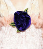 A model walks the runway wearing a pale pink ruffled mini dress with a large dark purple rose at the center of the bodice. The photo is taken during a fashion show with seated spectators on both sides.