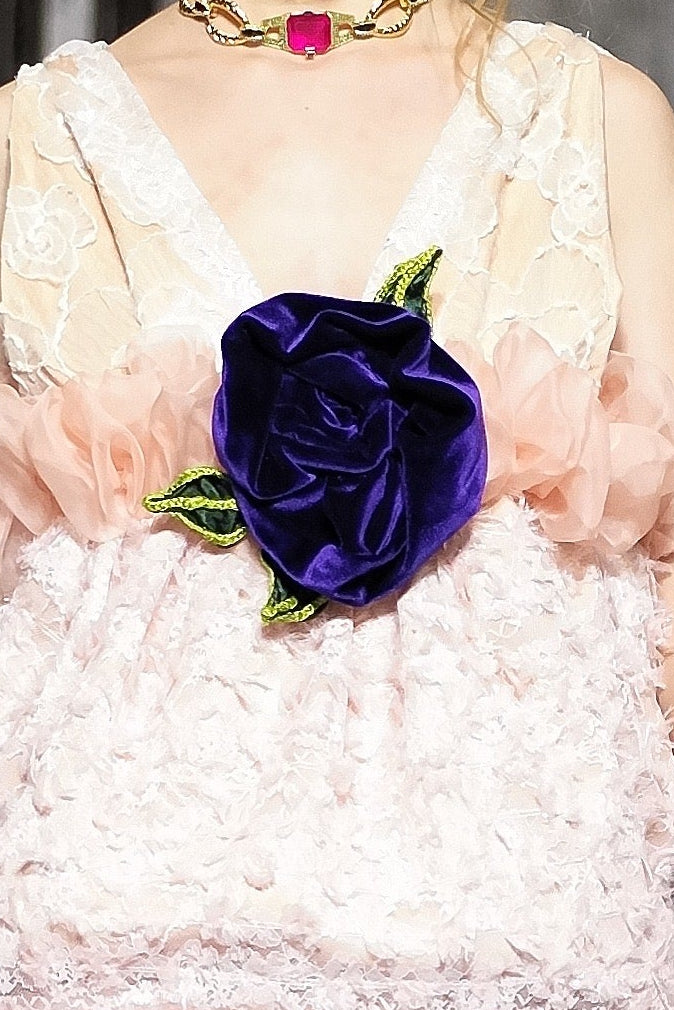 A model walks the runway wearing a pale pink ruffled mini dress with a large dark purple rose at the center of the bodice. The photo is taken during a fashion show with seated spectators on both sides.