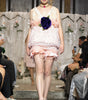 A model walks the runway wearing a pale pink ruffled mini dress with a large dark purple rose at the center of the bodice. The photo is taken during a fashion show with seated spectators on both sides.