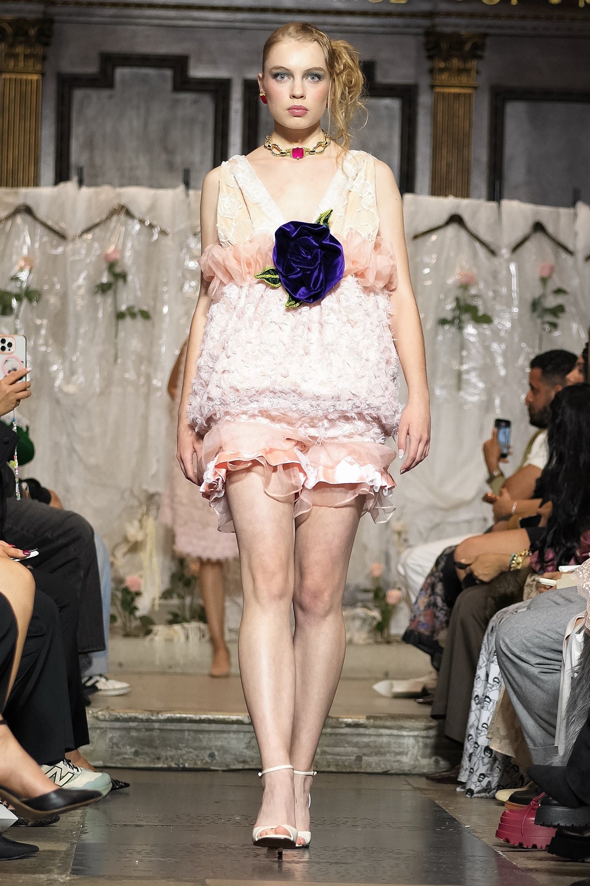 A model walks the runway wearing a pale pink ruffled mini dress with a large dark purple rose at the center of the bodice. The photo is taken during a fashion show with seated spectators on both sides.