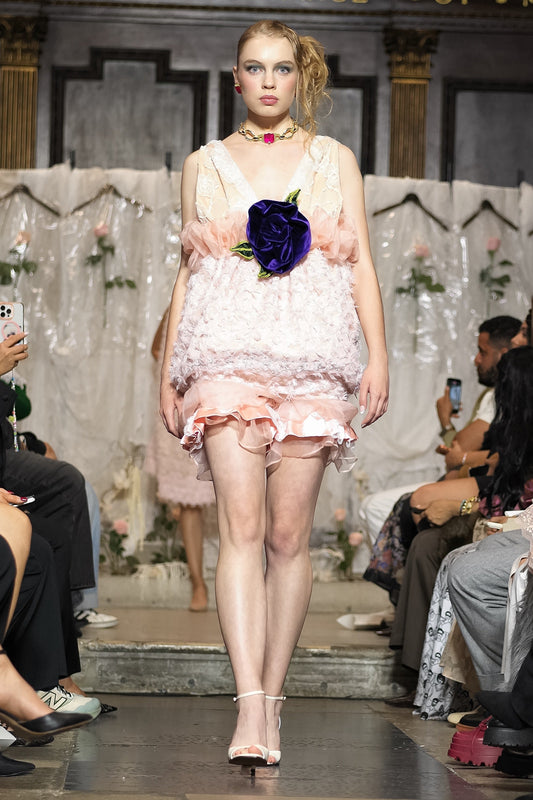 A model walks the runway wearing a pale pink ruffled mini dress with a large dark purple rose at the center of the bodice. The photo is taken during a fashion show with seated spectators on both sides.
